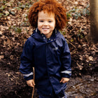 A young child smiling and wearing a Rainy Day Waterproof Jacket Navy in a woodland setting.